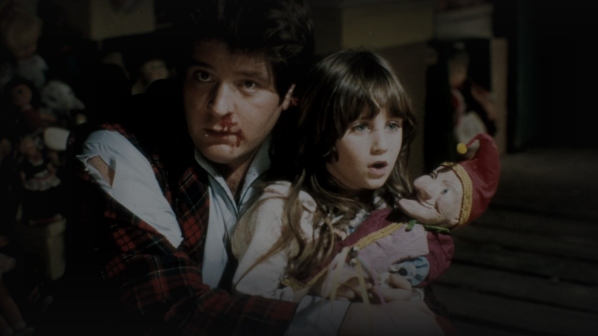 Still from Dolls (1987) — tmdb backdrop (ralph and judy with mr. punch, surrounded by dolls), source luma 72.4, baked at factor 0.45, final mean brightness 27.5